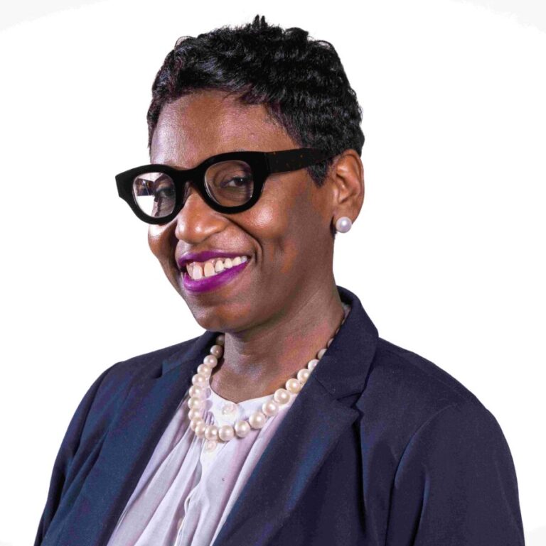 A person with short hair, wearing glasses, a pearl necklace, and a dark blazer over a light blouse, smiling at the camera against a white background.