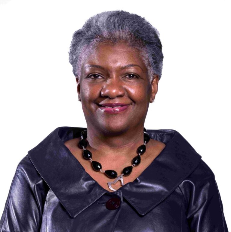 A smiling woman with short gray hair stands against a white background. She is wearing a black leather jacket with large buttons and a black beaded necklace.