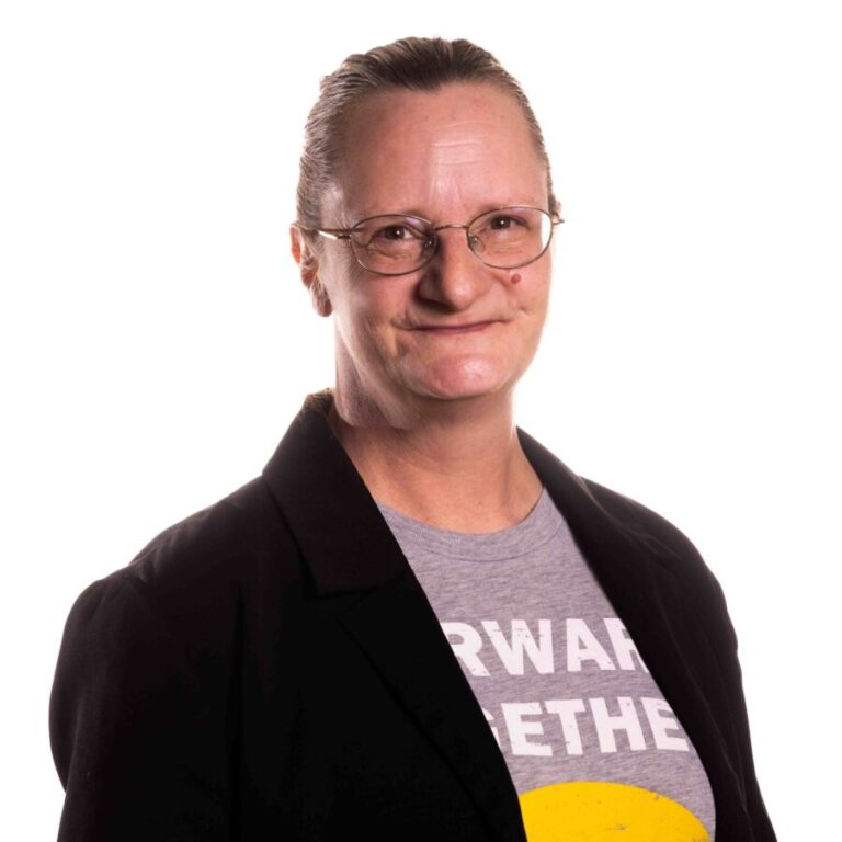 A person with light skin, brown hair pulled back, and glasses smiles softly at the camera. They are wearing a black blazer over a gray shirt with the partially visible words 