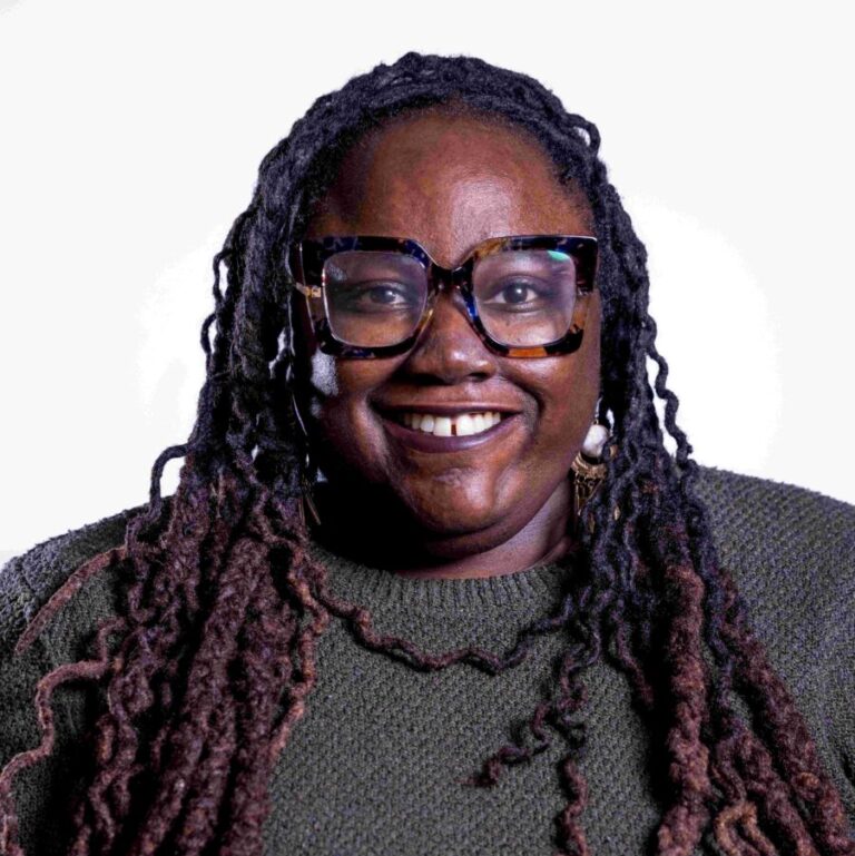 A person with long braided hair and large tortoiseshell glasses smiles while looking into the camera. They are wearing a green sweater and have large earrings. The background is plain white.