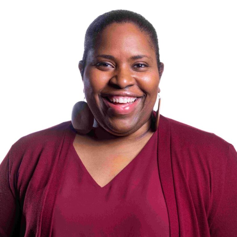 A person is standing against a white background, smiling directly at the camera. They are wearing a burgundy cardigan, matching top, and large round wooden earrings.