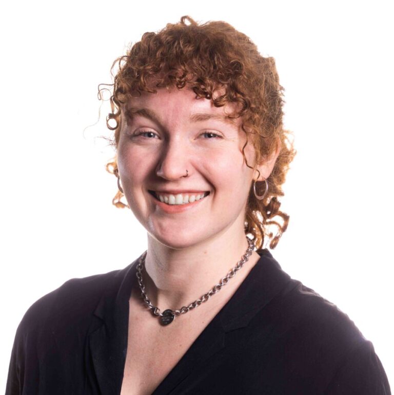 A person with curly brown hair is smiling at the camera. They are wearing a black top, a necklace, and small hoop earrings. The background is white and the lighting is bright.