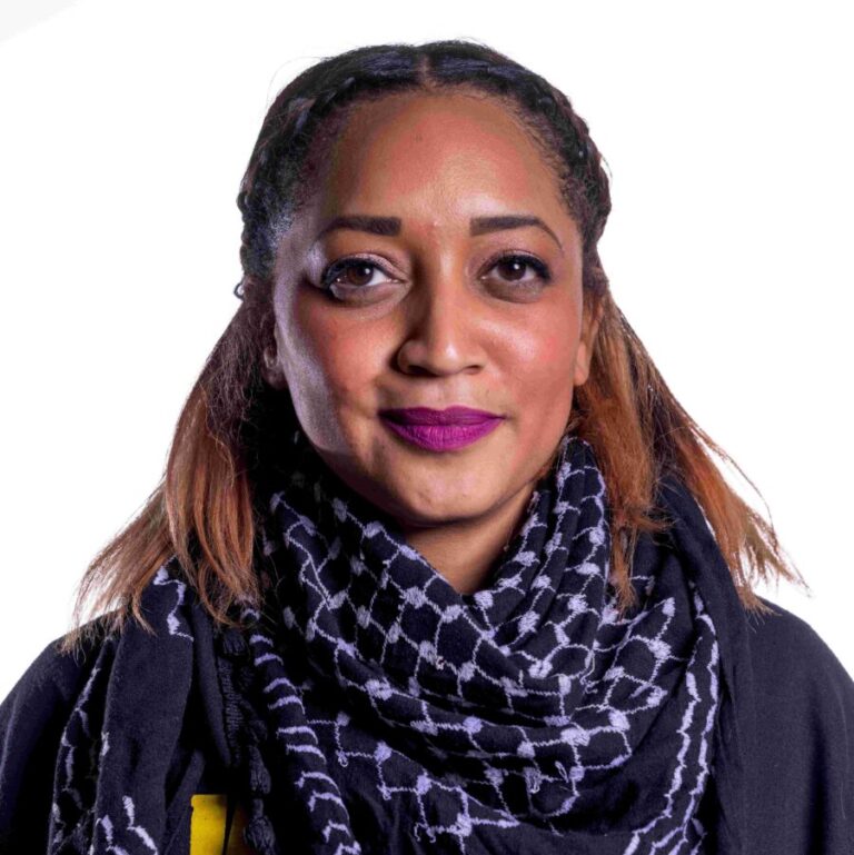 A person with long, dark braided hair wearing a black and white patterned scarf and a black shirt with yellow text stands against a plain white background. They have vibrant purple lipstick and smile slightly at the camera.