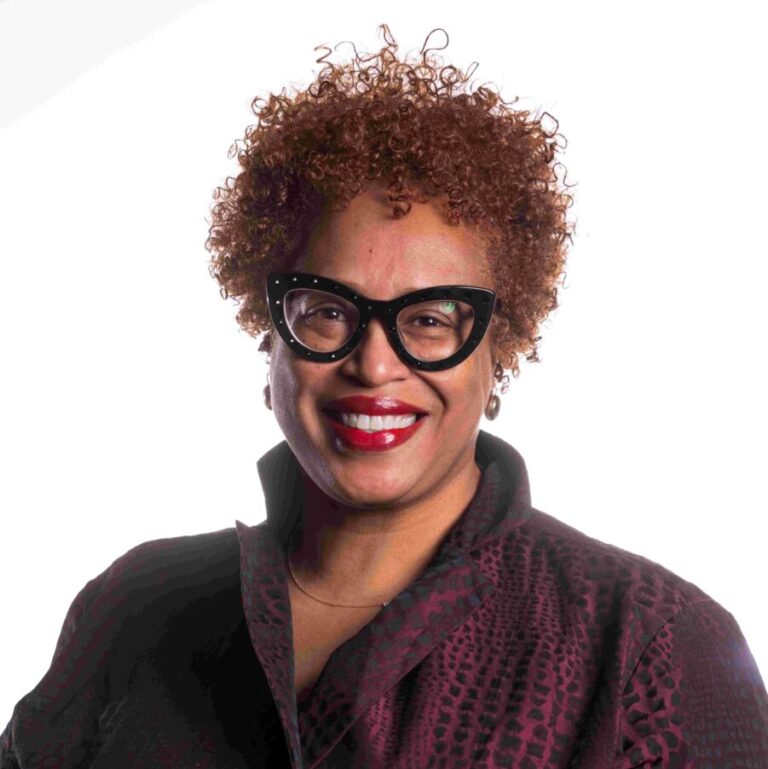 A person with short curly hair and black-framed glasses smiles while wearing a dark, textured jacket. The background is plain white, highlighting the subject's warm, welcoming expression.