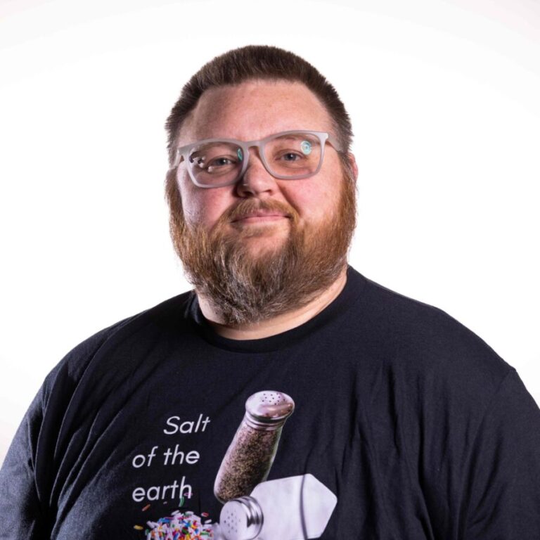 A person with a beard and glasses is wearing a black t-shirt. The shirt features an image of a salt shaker pouring colorful sprinkles instead of salt, with the text 