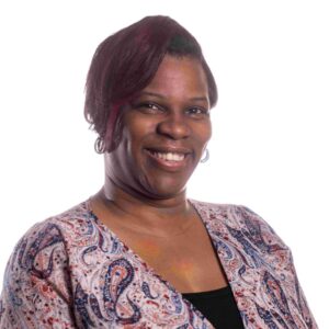 A smiling person with short, dark hair stands against a white background. They are wearing a long-sleeved, patterned top with paisley designs in blue, red, and white tones. The person looks at the camera with a friendly expression.