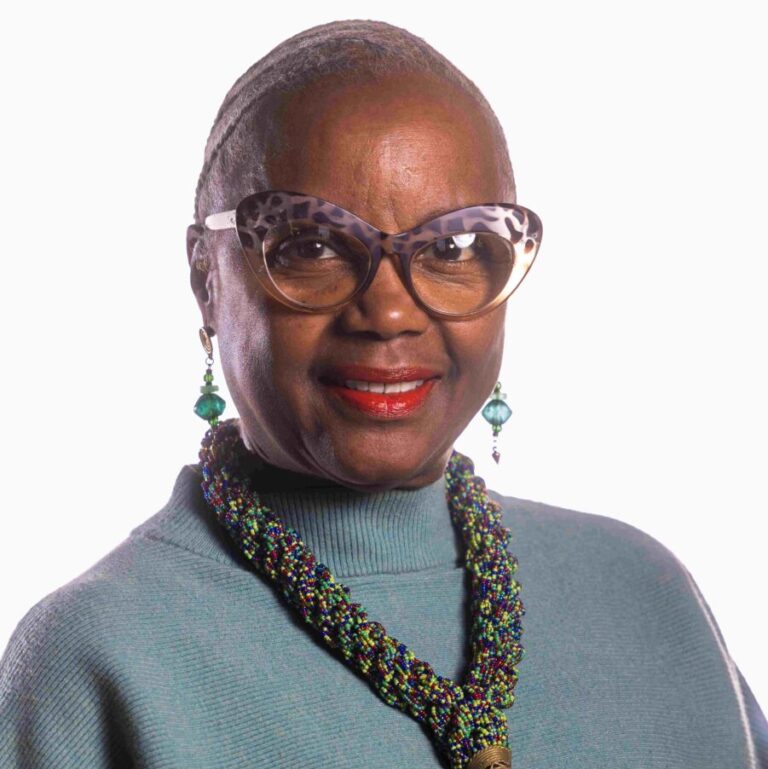 A woman with a shaved head, wearing large, round glasses, a teal long-sleeve top, and an intricately beaded necklace with a red pendant, smiles against a plain background. She also has teal earrings that match her top.