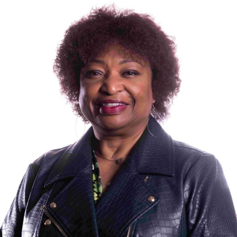 A woman with short, curly hair smiles at the camera. She is wearing a dark, textured jacket over a green patterned top. The background is white, and her posture is relaxed and confident.