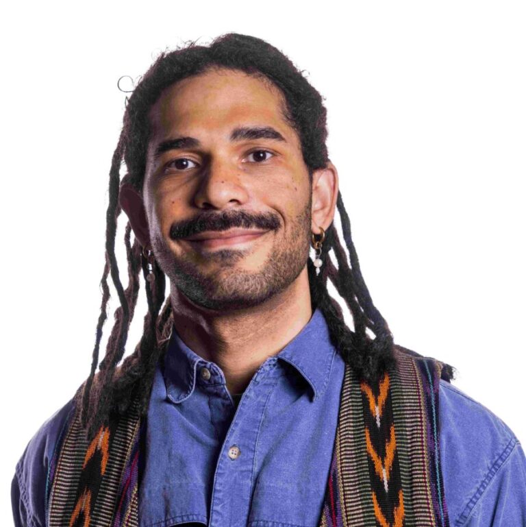 A person with long braided hair and a mustache smiles at the camera. They are wearing a blue button-up shirt and a patterned vest. The background is plain white.