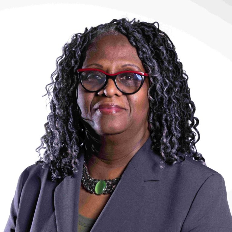 A woman with dark curly hair wearing red-rimmed glasses and a gray blazer stands against a white background. She is also wearing a green necklace and a green top. She is looking at the camera with a slight smile.
