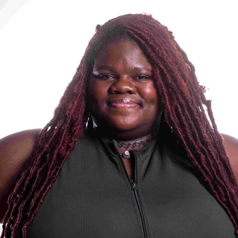 A person with long, braided reddish-brown hair smiles at the camera. They are wearing a dark sleeveless top with a zipper in the front and a choker necklace. The background is plain white.