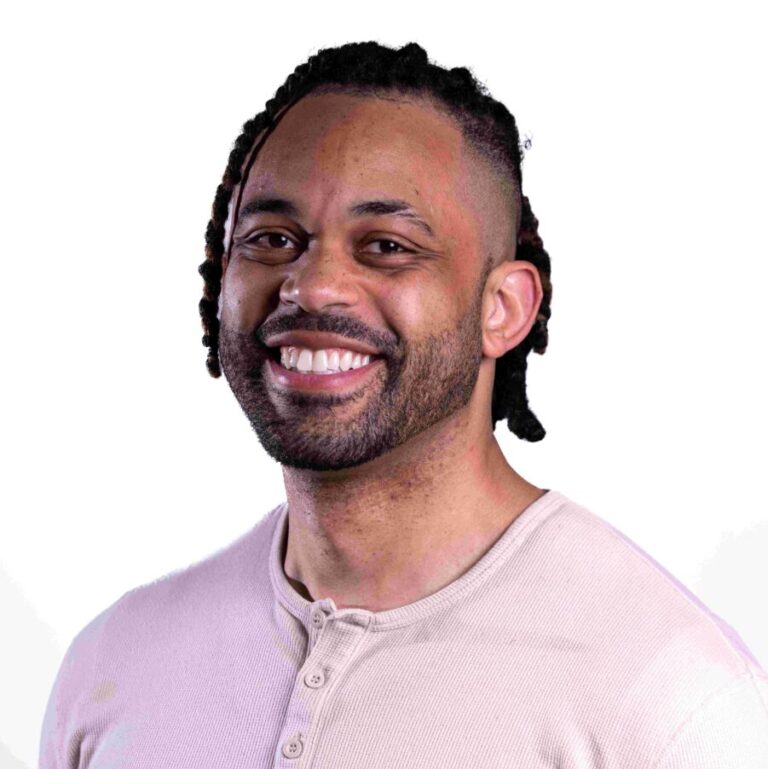 A smiling man with short dreadlocks and a neatly trimmed beard, wearing a light beige henley shirt. He is standing against a plain white background.
