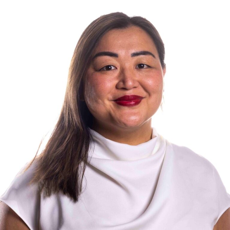 A woman with long, straight hair and red lipstick wears a white blouse with a draped collar. She has a slight smile and stands in front of a plain white background.