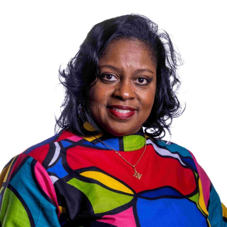 A woman with curled black hair smiles at the camera. She wears a vibrant, multicolored top with large abstract patterns in blue, red, green, and yellow. She also has a gold necklace with a pendant. The background is plain white.