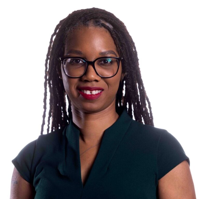 A person with long, braided hair, wearing black framed glasses, and a dark green short-sleeved top stands facing the camera. They are smiling and have a neutral, plain white background.