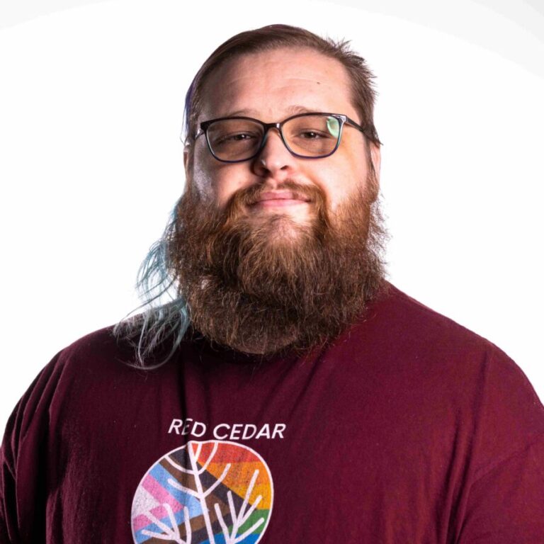 A person with long hair and a full beard smiles at the camera. They are wearing glasses and a maroon T-shirt that reads 