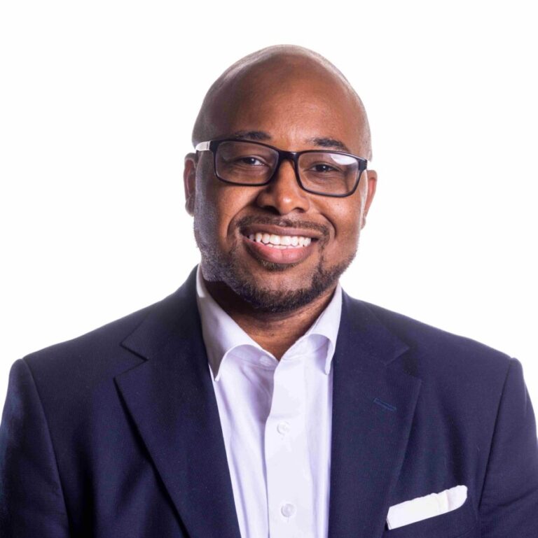 A person wearing a dark blue blazer, white dress shirt, and glasses is smiling at the camera. They have short hair and a neatly trimmed beard. The background is a plain white.
