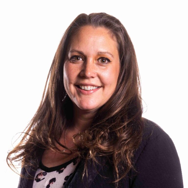 A woman with long brown hair and wearing a dark blazer over a light-colored patterned top smiles at the camera against a plain white background.