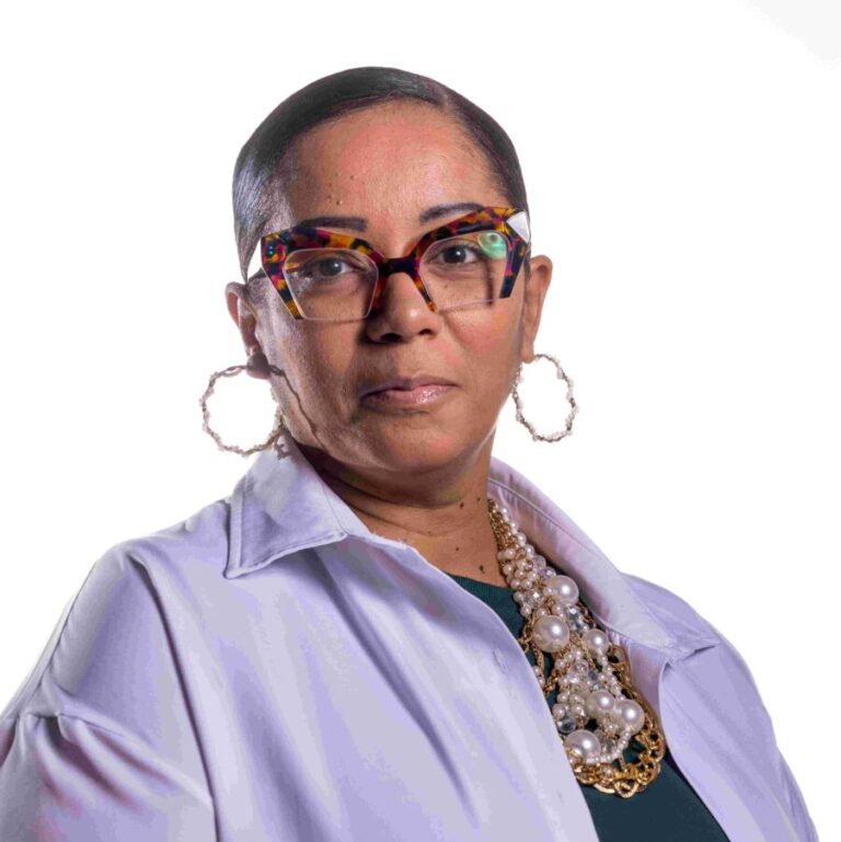 A person wearing colorful glasses, large hoop earrings, and a bold pearl necklace is shown from the chest up. They are dressed in a white collared shirt over a dark top, set against a plain white background. They have a serious expression and short, neatly styled hair.