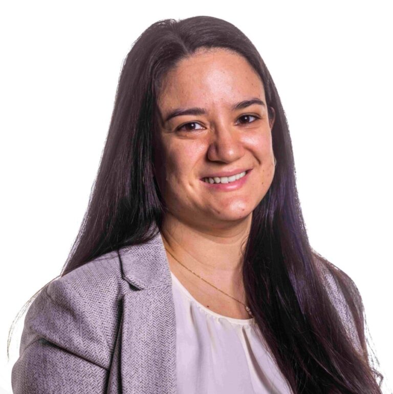 A person with long dark hair smiles at the camera. They are wearing a light-colored blazer over a white top. The background is plain and white.