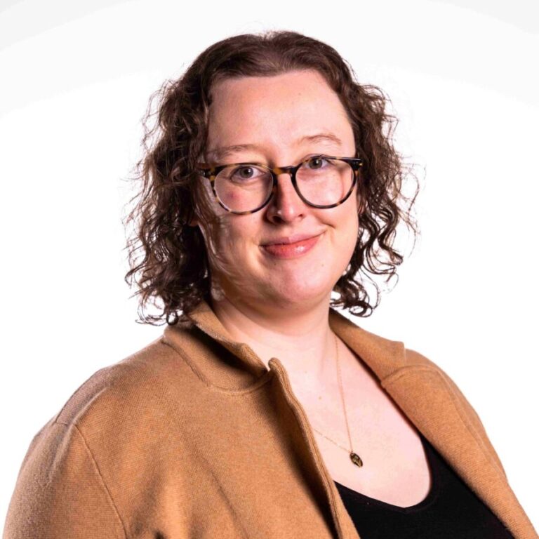 A person with shoulder-length curly hair, wearing glasses, and dressed in a brown jacket and black top, smiles slightly against a plain white background.