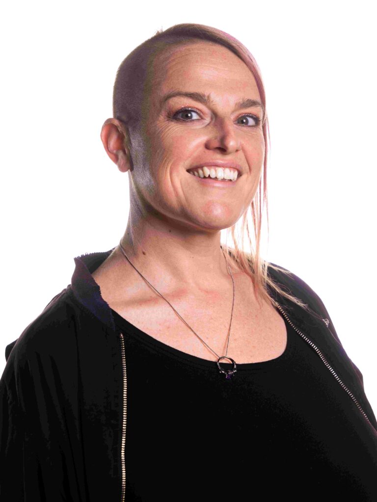 A smiling person with a unique hairstyle, including a shaved portion and a longer section swept to the side. They are wearing a black top and jacket, and a pendant necklace, posed against a plain white background.