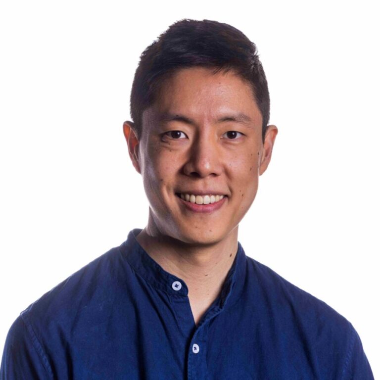 A person with short black hair wearing a dark blue button-up shirt is smiling at the camera against a white background.