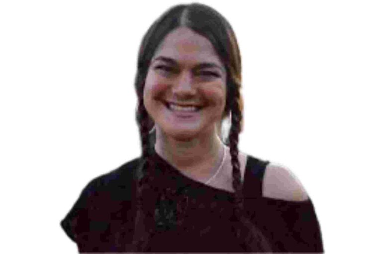 A young woman with long brown hair styled in two braids smiles warmly. She is wearing a black off-the-shoulder top and has a necklace around her neck. The background is plain white.