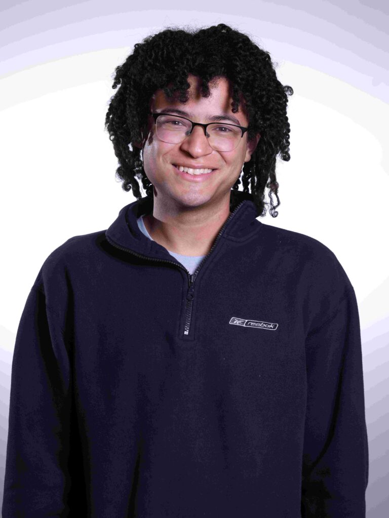 A smiling person with curly hair wearing glasses and a dark-colored zip-up sweater stands in front of a white and light purple gradient background.