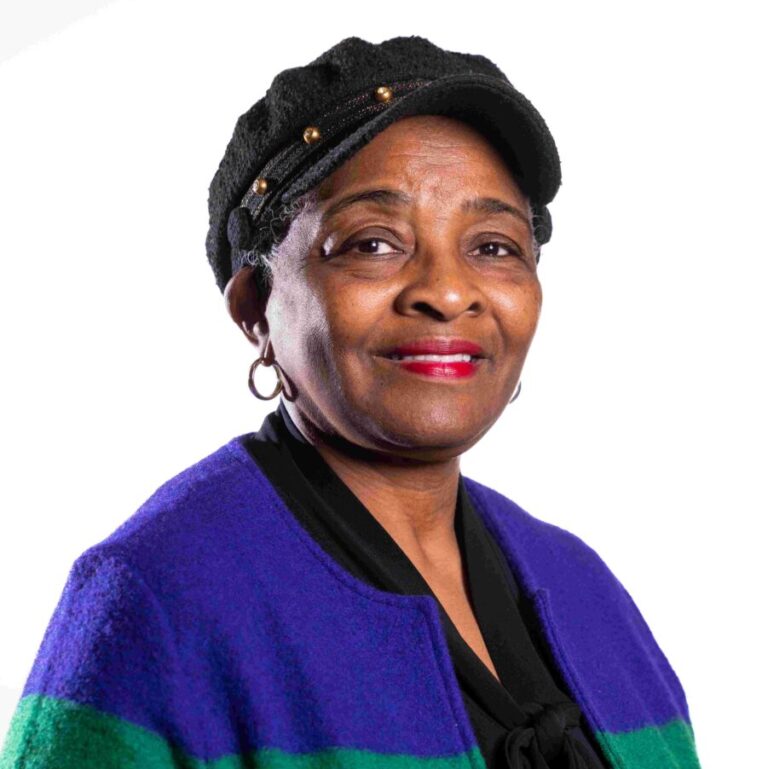 A woman is standing against a white background. She is wearing a black newsboy cap, large hoop earrings, and a colorful jacket with purple, green, and black sections. She has short hair and is smiling gently with red lipstick.