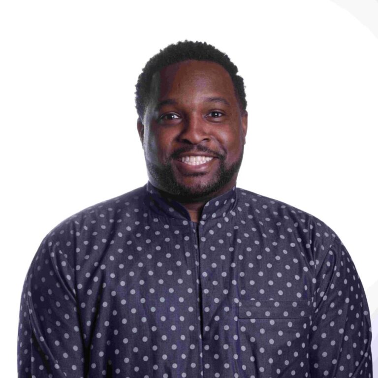 A smiling man with short curly hair and a beard is wearing a dark shirt with white polka dots. He stands in front of a plain white background.
