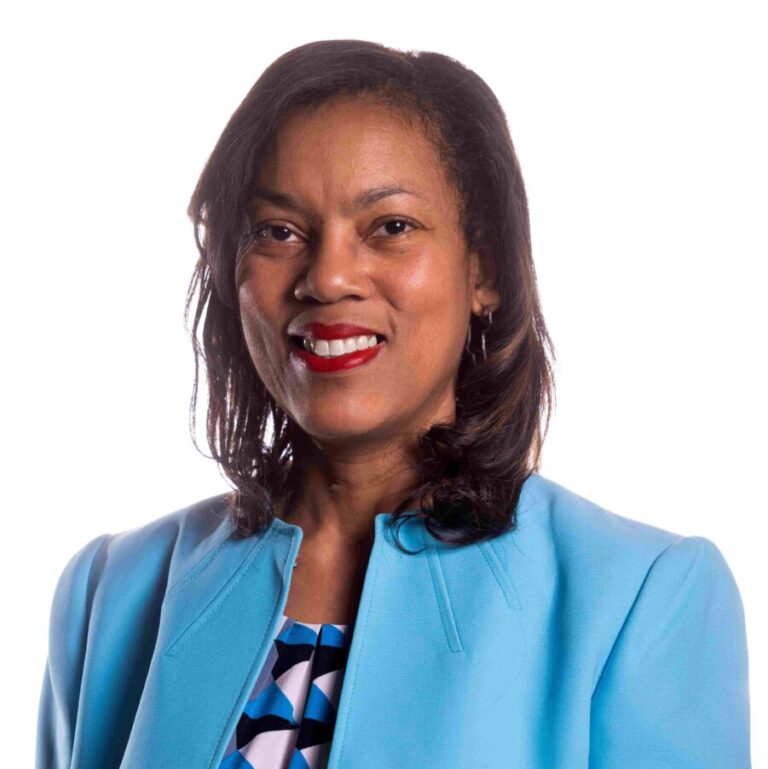A woman with shoulder-length dark hair smiles while looking at the camera. She is wearing a light blue blazer over a patterned shirt. The background is plain white.