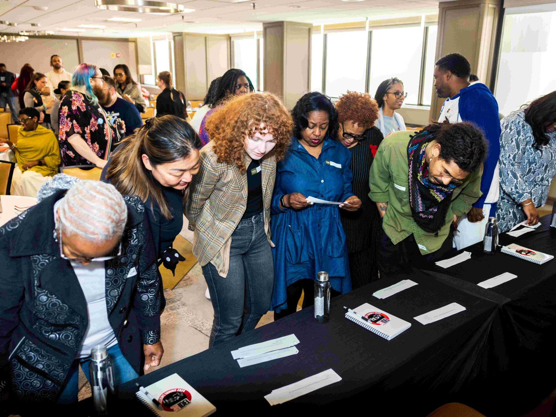 A diverse group of people leans over a table, engaging with materials displayed on it. They appear to be reading papers or examining items. The room is filled with natural light from large windows, and more people are gathered in the background.