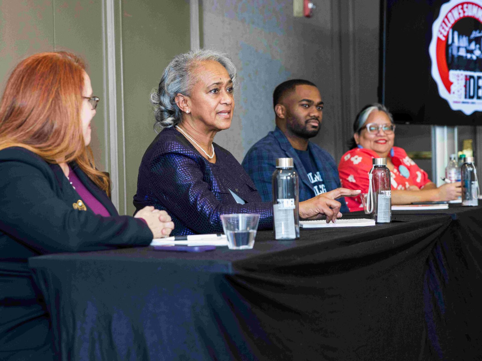 Four individuals are seated at a table in a panel discussion. The table is covered with a black cloth and has water bottles and papers. A logo with the text "IDEA" is visible in the background. The participants are engaged in conversation.