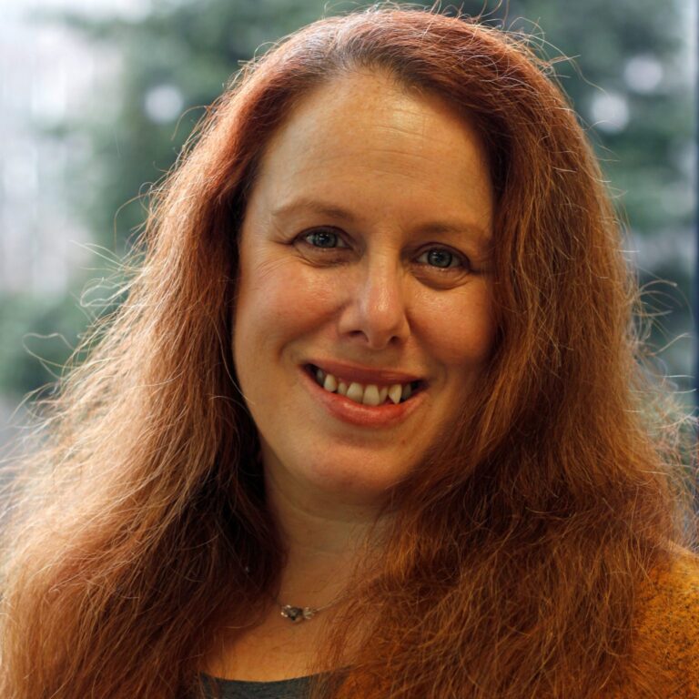 A woman with long, wavy red hair and a friendly smile is pictured in front of a blurred outdoor background. She is wearing a necklace and a grey top.