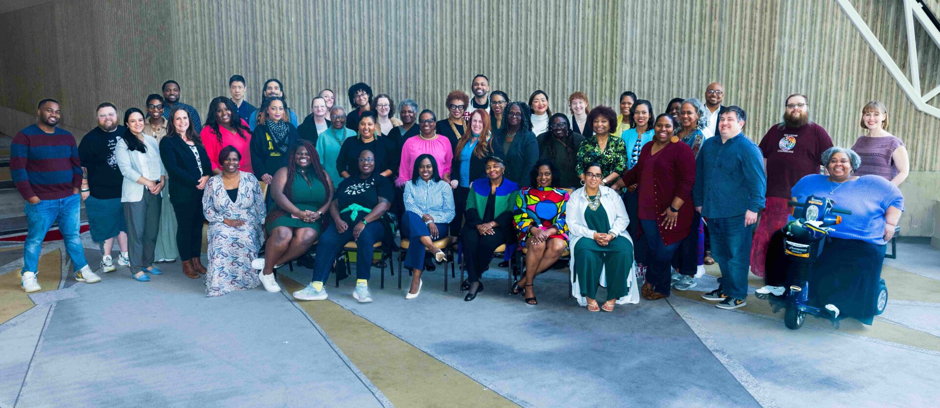 A diverse group of individuals poses for a group photo in an indoor setting. Most of them are standing, while some are seated in front. The group includes people of various ages, genders, and ethnicities, and one individual in a motorized wheelchair is present on the right.