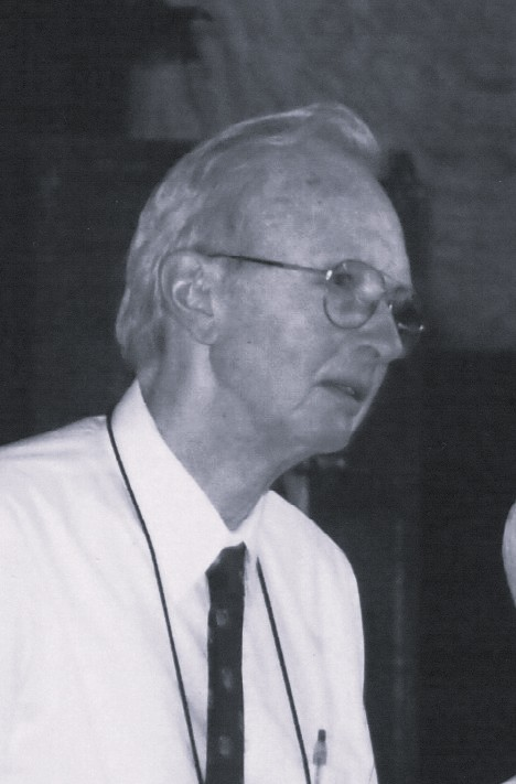 An older man with glasses and thinning light hair, wearing a white shirt, dark tie, and lanyard, stands indoors in a black-and-white photograph.