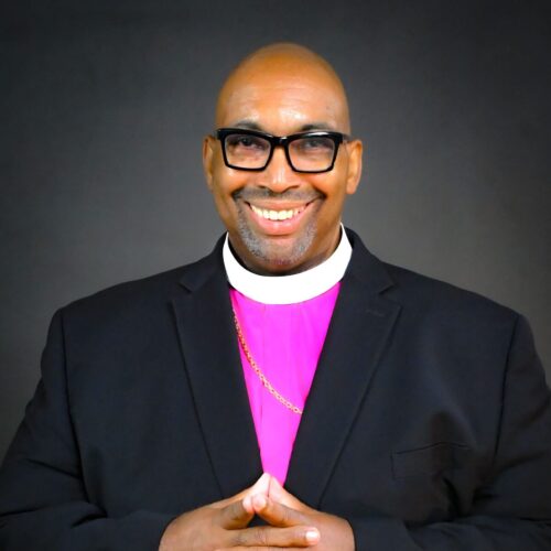 A man wearing glasses and a clerical collar over a magenta shirt and black suit embraces the camera with a warm smile. His hands are gently folded together against a plain dark background.