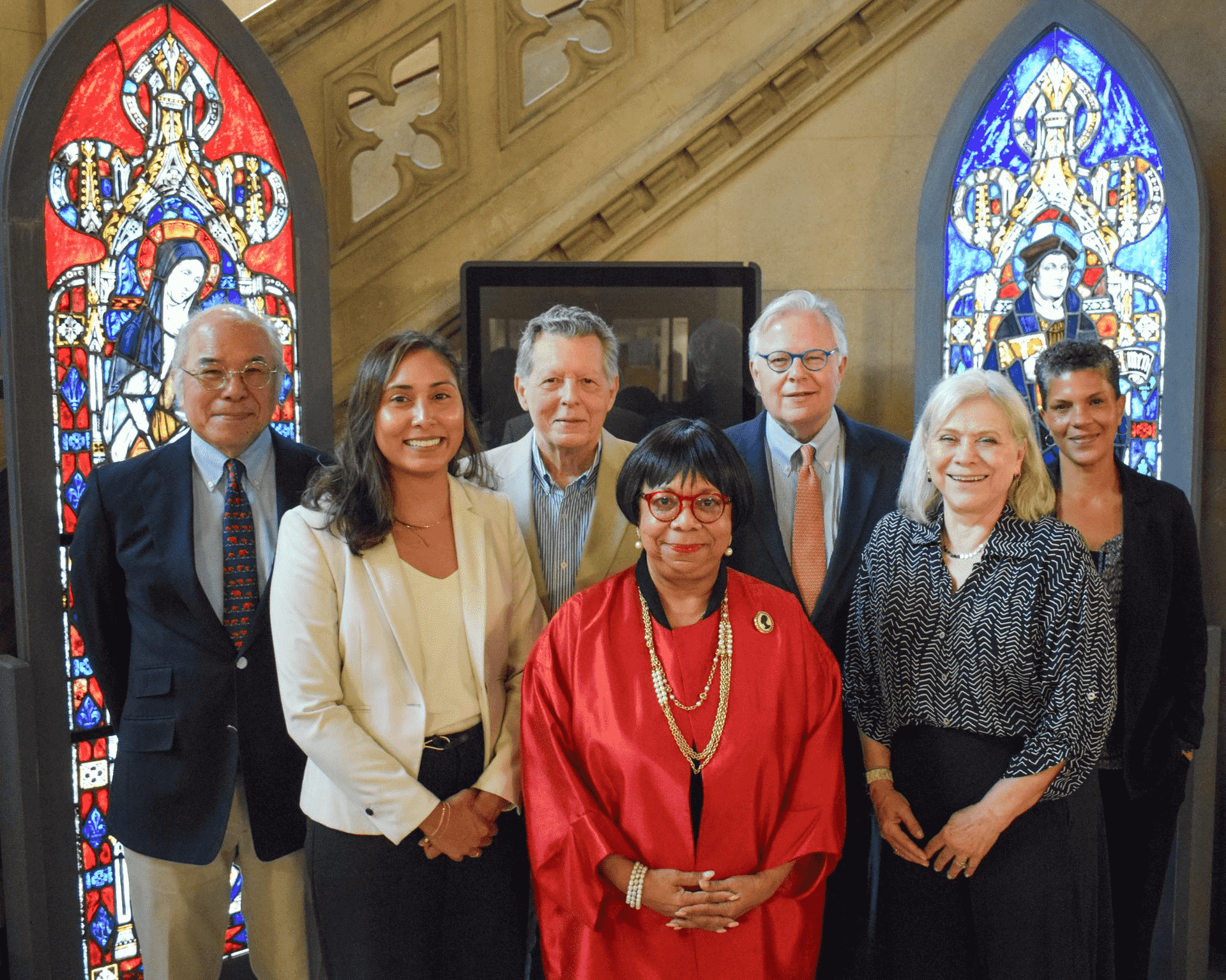 Seven people pose for a group photo indoors, standing in front of two colorful stained glass windows with religious imagery and a stone staircase in the background.