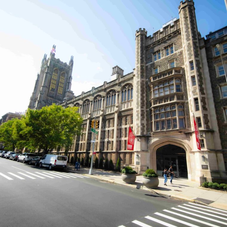 A large, historic stone university building with gothic architecture stands along a tree-lined city street; people walk near the entrance, and red banners hang by the doorway.