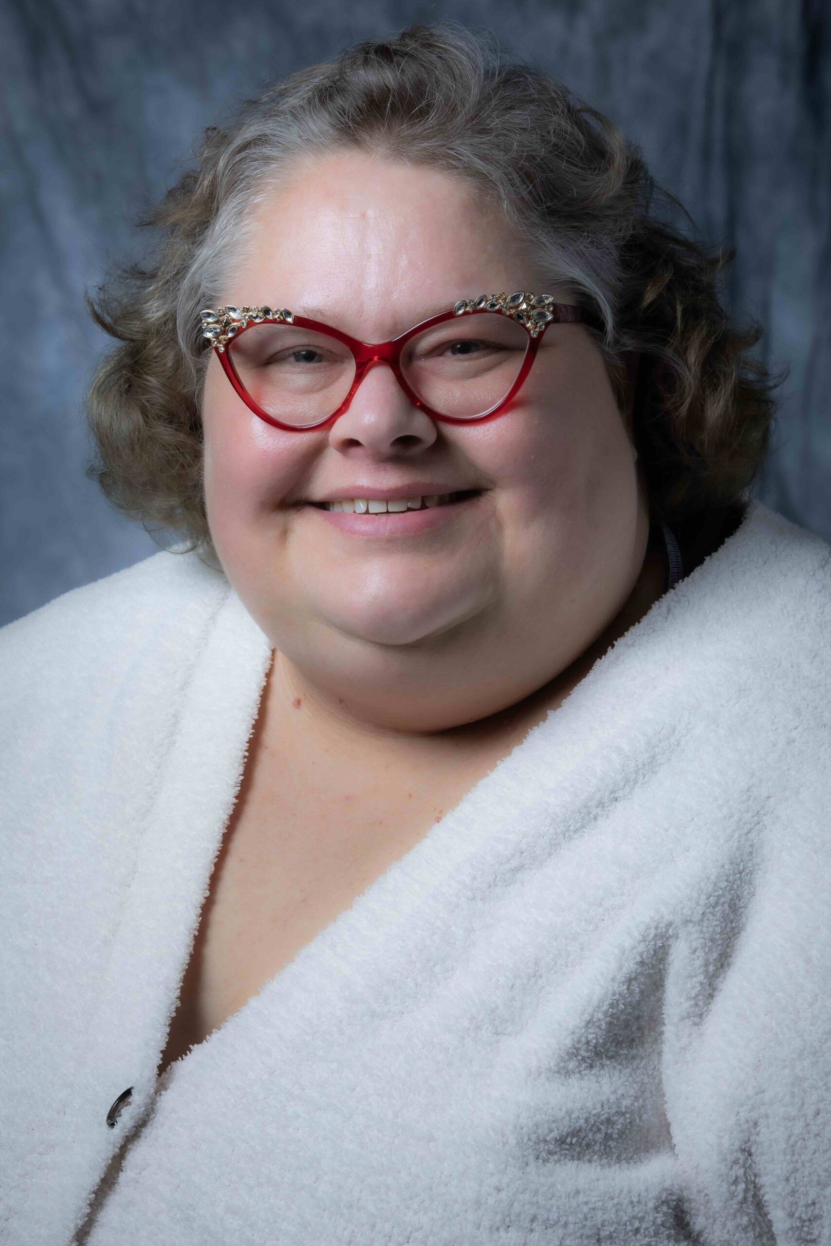 A person with short curly hair smiles warmly at the camera. They are wearing red glasses and a white textured top, in front of a gray background.