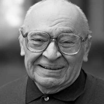 An elderly man with glasses and a smile, wearing a dark collared shirt, is shown in a black and white portrait. The background is blurred, highlighting his face.