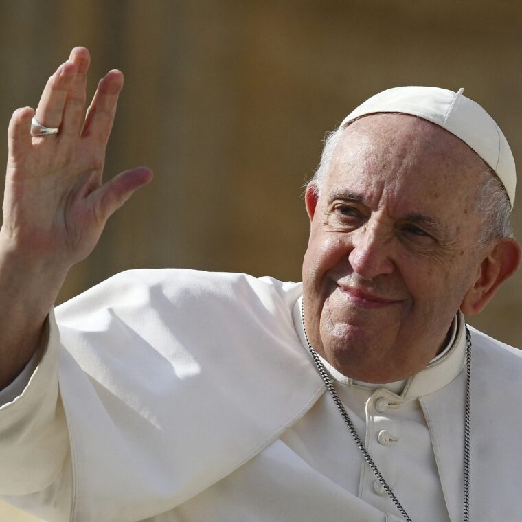 An elderly man in white religious robes and a white cap smiles and waves with his right hand. He is outdoors, sunlight illuminating his face—a warm scene often seen in update about Pope Francis or Pope Francis news.