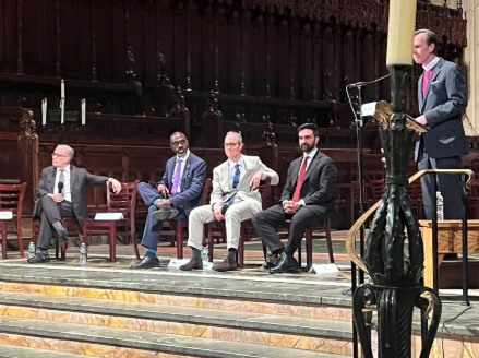 Five men in suits participate in a panel discussion at the front of a church, with four seated on chairs and one standing at a lectern speaking into a microphone. Candles and dark wooden panels are visible in the background.