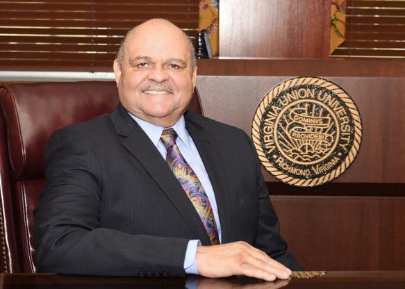 A man in a suit sits at a desk, smiling. Behind him is a seal that reads "Virginia Union University, Richmond, Virginia." The setting appears to be an office or formal environment.