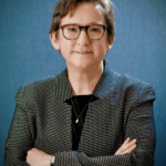 A person with short brown hair and glasses is wearing a patterned blazer, a black top, and a necklace, standing with arms crossed in front of a blue background.