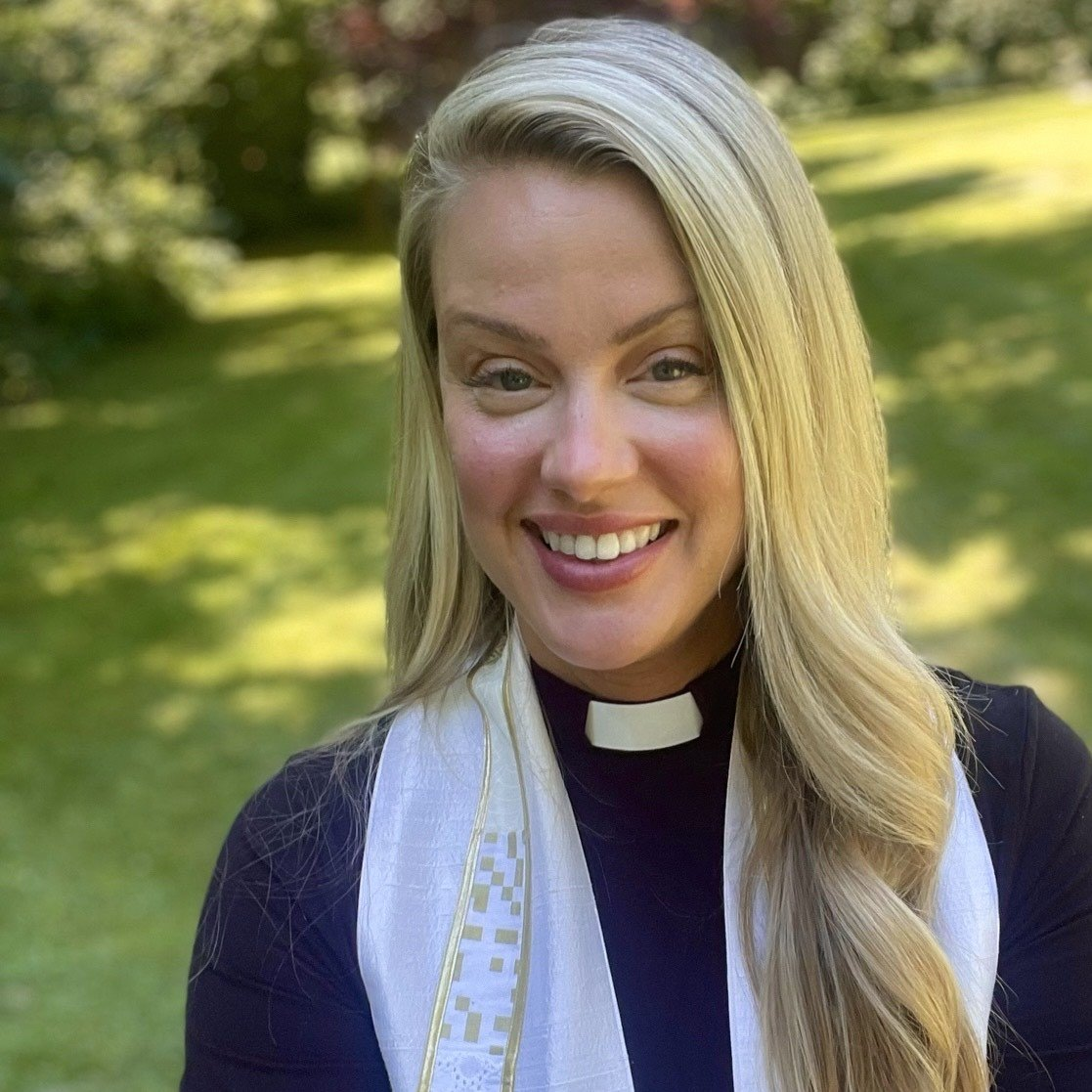 Smiling person with long blonde hair and a clerical collar stands outside in a green garden. They are wearing a black top and a white stole with gold details. The background is lush and sunlit.