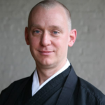 A person with a shaved head wearing traditional black and white robes stands in front of a neutral, light-colored background, looking at the camera with a slight smile.