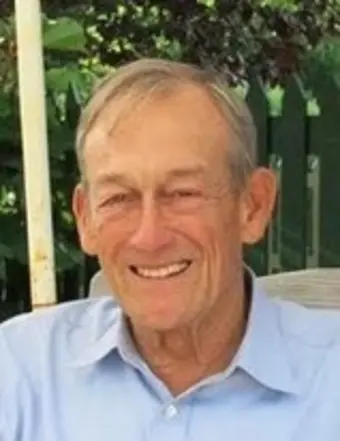 An older man with short gray hair smiles while sitting outdoors. He is wearing a light blue collared shirt, and greenery is visible in the background.
