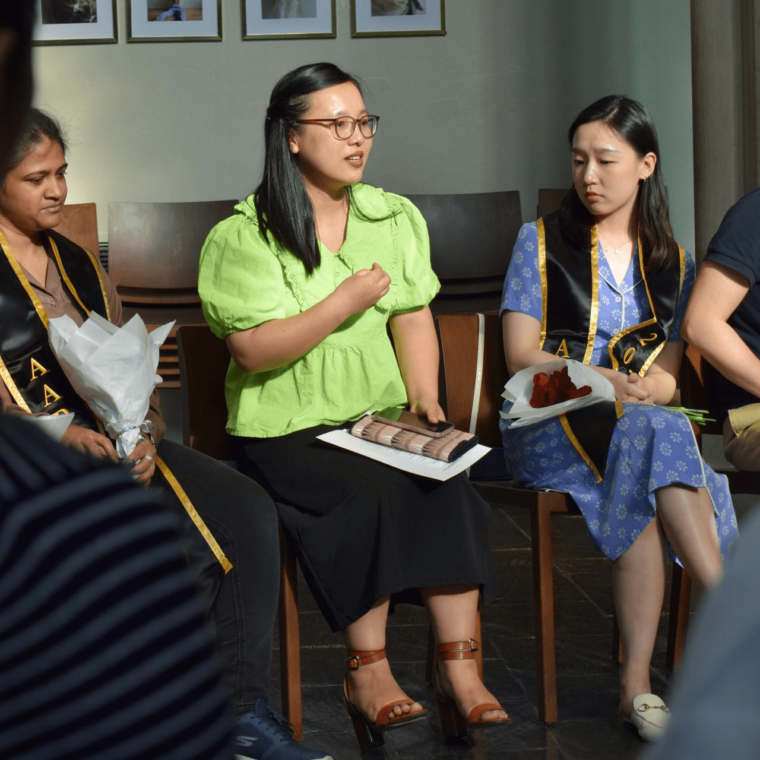A group of people sitting on chairs in a semicircle. One person wearing glasses and a green blouse is speaking, holding a book. Others listen attentively. Some wear graduation stoles. Framed photos hang on the wall in the background.